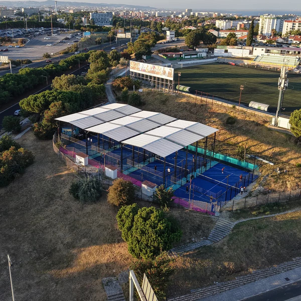 O clube visto de cima ao fim da tarde, entre pinheiros, com o campo de futebol vizinho e os bairros de Lisboa ao fundo.
