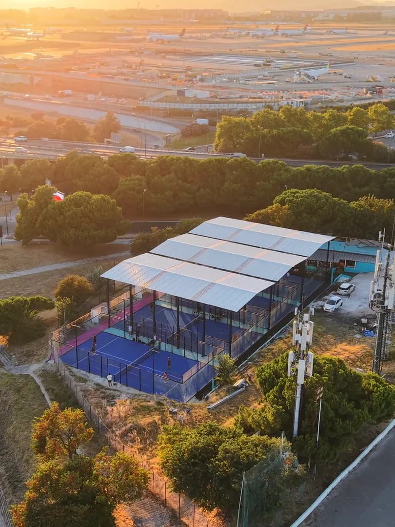 O Padel do Relógio visto de drone ao pôr do sol: os campos azuis sob a cobertura branca do clube, com as pistas do aeroporto de Lisboa e a serra douradas pelo sol ao fundo.